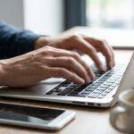 Die Aufnahme zeigt eine Person, die auf einer Tastatur eines Laptops tippt, während ein Smartphone und eine Tasse Kaffee auf dem Tisch liegen.