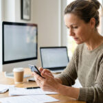 A local business owner carefully checks their company website on a smartphone at a desk, with a computer in the background.